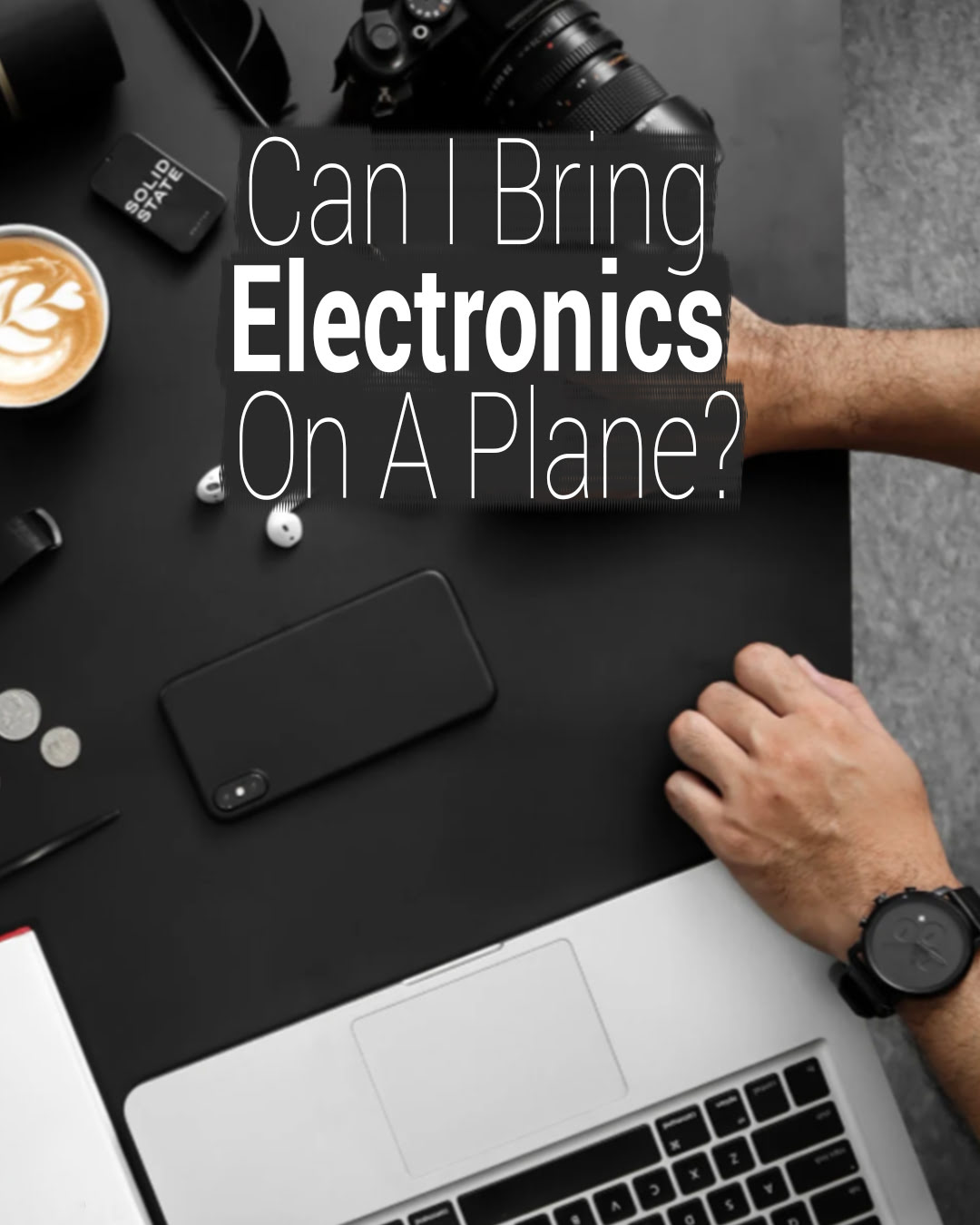 Flat lay of a workspace with electronics including a laptop, smartphone, camera, wireless earbuds, a smartwatch, and a cup of latte art, with the text Can I Bring Electronics On A Plane