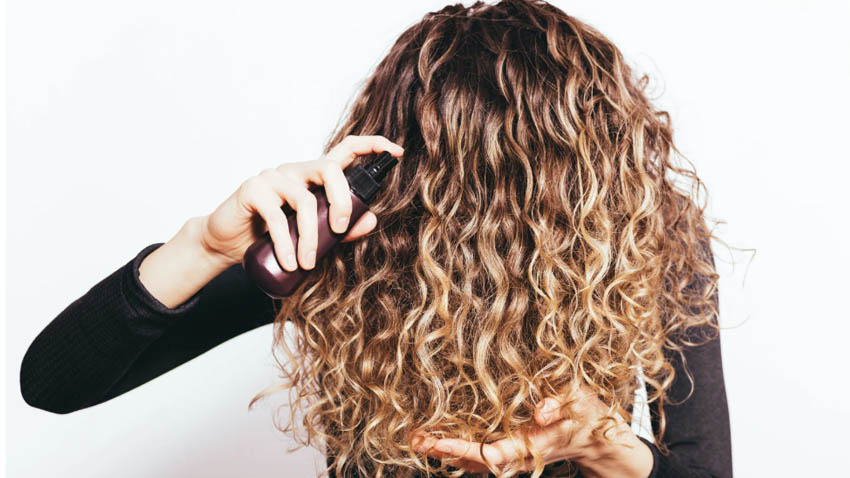 Woman spraying styling mousse on curly hair, demonstrating common use of hair products for air travel articles.